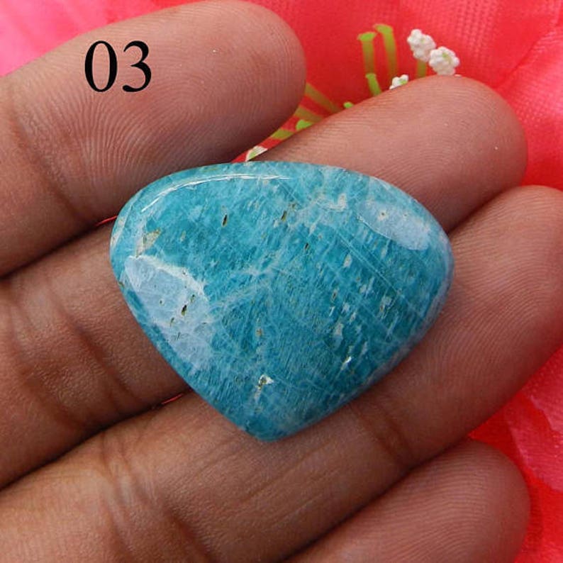 Puede incluir: Un cabuj&oacute;n de amazonita verde azulado con vetas blancas y forma de coraz&oacute;n. La piedra se sostiene en una mano.