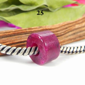 Puede incluir: Primer plano de una cuenta de piedra preciosa rosa en una cadena de plata. La cuenta es redonda y tiene un agujero en el centro. La cuenta está rodeada por una superficie de madera.