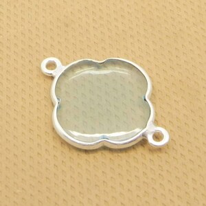 Pode incluir: Pingente de metal prateado com um centro de vidro transparente, em forma de trevo de quatro folhas. O pingente tem dois laços na parte superior para prender uma corrente ou um cordão.