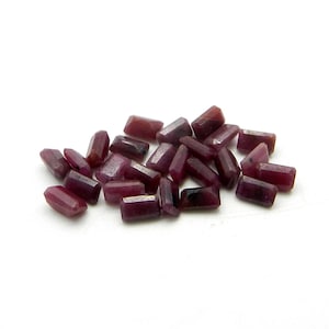 May include: A pile of small, rectangular ruby gemstones. The gems are a deep red-purple colour with faceted edges, scattered on a white surface. These gemstones are likely for jewellery making or crafting.