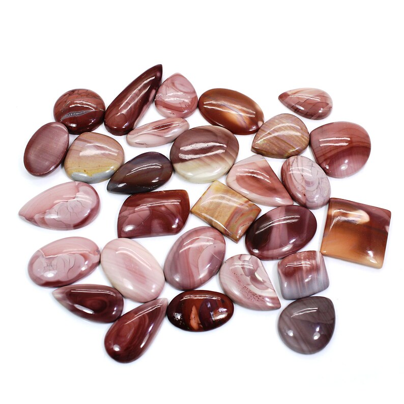 May include: A collection of 20 polished, natural, red, brown, and pink gemstone cabochons. The cabochons are various shapes and sizes, including oval, teardrop, and rectangular.