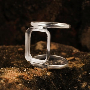 May include: A silver ring with a distinctive design. The ring has a rectangular central element, with bands at the top and bottom. The ring is made of a shiny, reflective metal and is set against a textured, dark background.