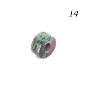 May include: A green and pink gemstone bead with a hole in the center. The bead is oval shaped and has a rough texture. The bead is labeled "14".