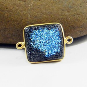 Pode incluir: Um pingente quadrado dourado com um centro brilhante azul escuro e azul claro. O pingente tem dois pequenos laços em cada lado para prender a uma corrente ou outro componente de joalheria.