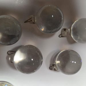 May include: Five clear, spherical glass ornaments, each featuring a small, integrated metal loop at the top for hanging. The smooth, transparent glass spheres are arranged in a cluster, reflecting light on their polished, rounded surfaces.