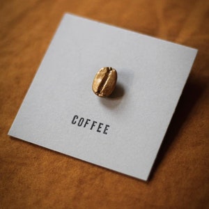 Può includere: Un chicco di caffè dorato è appoggiato su un biglietto quadrato grigio. La parola "COFFEE" è stampata in nero sotto il chicco. Il biglietto è posto su uno sfondo marrone strutturato.