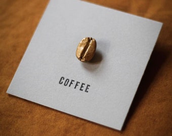 May include: A golden coffee bean rests on a gray square card. The word "COFFEE" is printed in black below the bean. The card is set against a brown, textured background.