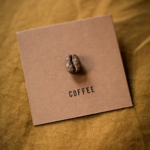 Può includere: Un singolo chicco di caffè tostato è appoggiato su un cartoncino quadrato marrone. La parola "COFFEE" è stampata in nero, centrata sotto il chicco. Il cartoncino è posto su uno sfondo giallo dorato strutturato.