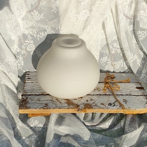 Op de afbeelding: Een matglazen bolvormige lampenkap. De lampenkap is rond met een brede basis en een kleinere opening aan de bovenkant. Hij staat op een verweerde, wit geschilderde houten plank, waaraan een touw is bevestigd. De achtergrond is een wit kanten gordijn.