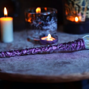 May include: A purple and silver wand with a clear glass orb at the end. The wand is lying on a wooden surface with candles and a glass jar in the background.
