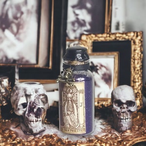 May include: A glass bottle filled with purple glitter and a skull charm. The bottle has a label with the text "High Priestess" and a tarot card image. The bottle is sitting on a gold tray with two white skulls.