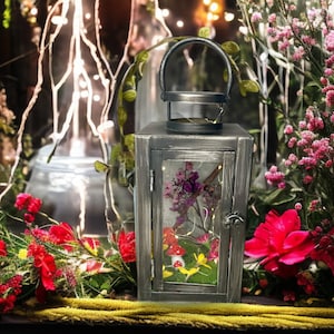May include: A decorative gray metal lantern with a handle, glass panels, and a hinged door. The lantern is filled with dried flowers, fairy lights, and miniature mushrooms. Red and pink flowers surround the lantern.