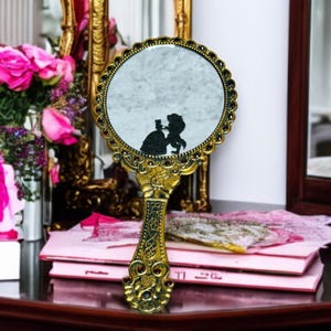 Peut inclure: Un miroir de main de style vintage avec un cadre et une poignée dorés ornés. Le miroir rond présente une silhouette noire d'un couple. Le miroir est sur une table avec des livres et des fleurs roses.