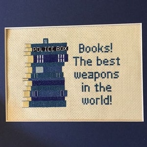 Pode incluir: Bordado de ponto cruz de uma caixa de polícia azul com o texto "Police Box" acima. O texto "Books! The best weapons in the world!" está bordado abaixo da caixa.