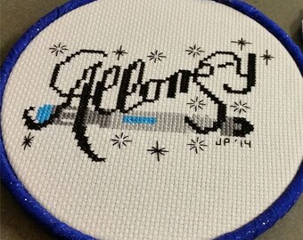 May include: A white embroidery hoop with a blue ribbon border. The hoop features a black and white cross-stitch design of the word "Galaxy" with a small blue and gray spaceship underneath. The design is surrounded by small white stars.