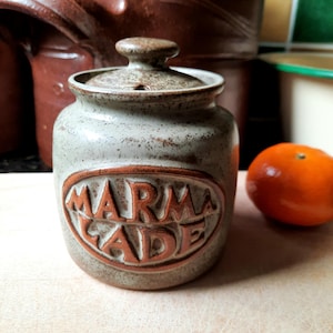 May include: A stoneware marmalade jar with a lid. The jar is pale grey with brown flecks and a brown oval with the word "MARMALADE" in relief. An orange fruit is to the right.