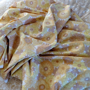 May include: A folded fabric featuring a repeating floral design in yellow, gold, and brown hues. The flowers are large and stylized, with a retro style. The fabric appears to be cotton or a similar material, with a soft drape, suitable for various projects.