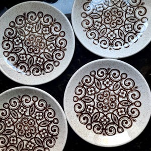 May include: Four white ceramic plates with a speckled finish. Each plate has a brown floral and swirl pattern in the centre. The plates are arranged in a square formation, showcasing the decorative design.
