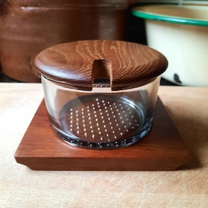 May include: A glass jar with a wooden lid and base. The lid has a small notch. The jar sits on a square wooden base. The jar has a perforated metal insert. The wood is a medium brown colour.