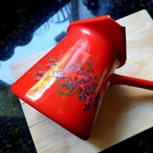 May include: A red enamel coffee pot with a long handle and a spout. The pot is decorated with a floral design in shades of purple, pink, and green. The pot is sitting on a wooden cutting board.