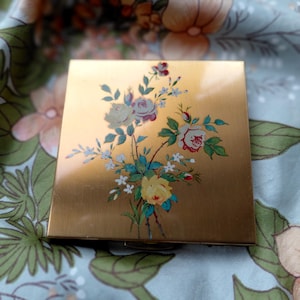 May include: A gold-coloured compact with a floral design. The square compact features a painted bouquet of roses in shades of red, yellow, and white, with green and blue leaves. The compact is closed and appears to be vintage.
