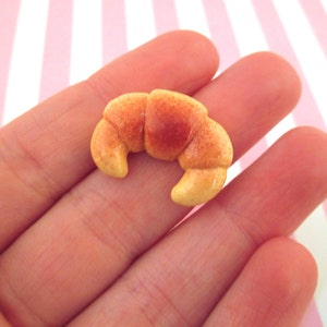 May include: A small, light brown croissant made of polymer clay. The croissant is about the size of a fingernail.