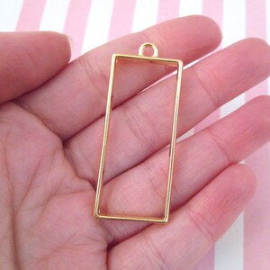 May include: Gold-toned metal pendant frame with a rectangular shape and a loop at the top for hanging. The frame is empty and ready for resin or other embellishments.