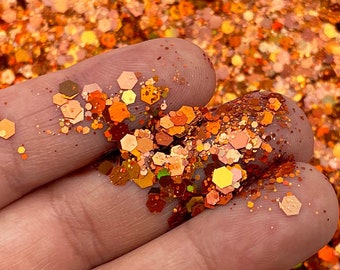 May include: A close-up of a pile of orange, gold, and copper glitter. The glitter is in various shapes and sizes, and it has a metallic sheen.