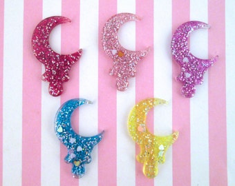May include: Five glitter crescent moon charms in different colors: pink, blue, yellow, light pink, and white. The charms are made of resin and have a glittery finish.