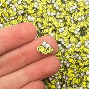 May include: A close-up of a hand holding a small cluster of yellow and black bee-shaped charms. The charms are made of polymer clay and have a glossy finish. The background is a large pile of similar charms.