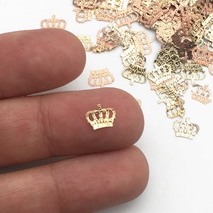 May include: A pile of gold, rose gold, and silver crown shaped metal embellishments. The crowns are scattered on a white surface.