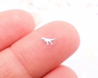 May include: A silver dinosaur cutout, approximately 1/2 inch in size, is held between two fingers. The dinosaur is facing right and has a long tail.