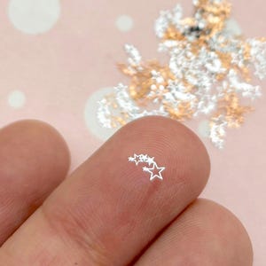 May include: Close-up of tiny silver star-shaped confetti. A few stars are on a fingertip, with a larger pile of silver and gold stars in the background. The confetti is scattered on a pink background with white polka dots.
