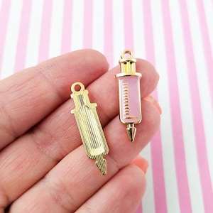 Puede incluir: Dos abalorios de metal dorado en forma de jeringa. Un abalorio es rosa con detalles dorados. El otro abalorio es dorado con textura acanalada.