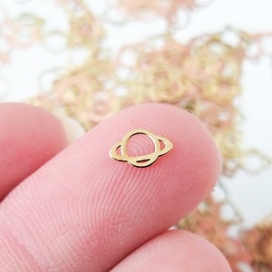 Op de afbeelding: Een gouden metalen bedel in de vorm van een planeet met een ring eromheen. De bedel is klein en delicaat en perfect om een ​​toetsje whimsy toe te voegen aan sieraden of knutselwerk.