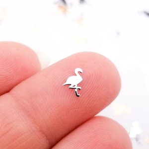 May include: A tiny, silver-colored flamingo charm. The charm is a miniature representation of the bird, with its long neck and legs. The flamingo is perched on a small, curved leg. The charm is held between two fingers.
