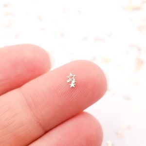 May include: A close-up of a fingertip holding a small cluster of silver star-shaped nail charms. The charms are scattered on a white surface.