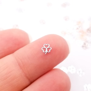 Könnte beinhalten: Ein kleiner silberner Glücksbringer in Form eines vierblättrigen Kleeblatts, ein kleiner Metall-Anhänger in Form eines Kleeblatts, der zwischen zwei Fingern gehalten wird.