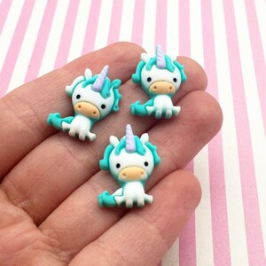 May include: Three turquoise and white unicorn charms with purple horns. The charms are small and have a cartoon-like appearance.