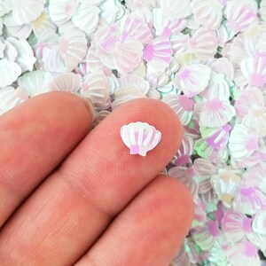 May include: A close-up of iridescent white seashell-shaped glitter. The glitter is scattered on a surface and a finger is holding a single piece of glitter.