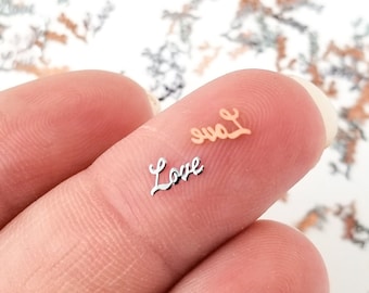 May include: Two small metal charms with the word "Love" written in a cursive font. The charms are silver and rose gold in color.