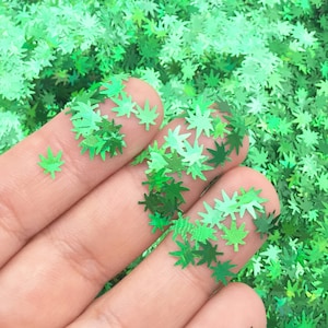 May include: Green glitter in the shape of marijuana leaves. The glitter is loose and scattered on a surface.