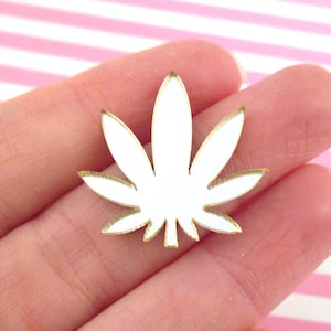 May include: White marijuana leaf cutout with a gold outline. The leaf is held in a hand against a pink and white striped background.