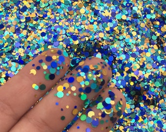 May include: A close-up of a hand holding a variety of blue, green, and gold glitter. The glitter is in the shape of small circles.