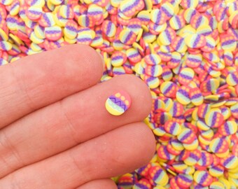 Pode incluir: Um close-up de uma mão segurando uma pequena confete de ovo de Páscoa colorida. A confete é rosa, amarela e roxa com um padrão de ziguezague branco. O fundo está cheio de muitas outras confete iguais.