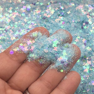 Può includere: Primo piano di glitter blu e bianco iridescente a forma di farfalla. Il glitter è in un mucchio e una mano ne tiene un po'.