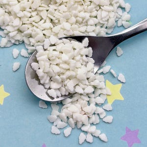 May include: A pile of small, white, irregularly shaped sprinkles next to a silver spoon. The sprinkles are on a light blue surface decorated with colorful star-shaped confetti.