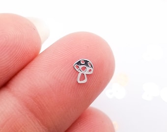 May include: A tiny silver mushroom charm with a black and white dotted cap is held between fingers. The charm is a miniature representation of a mushroom, with a detailed design. The background is white with scattered mushroom-shaped confetti.