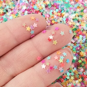 May include: A hand holding a variety of iridescent star-shaped glitter. The glitter is a mix of pink, blue, green, and yellow.