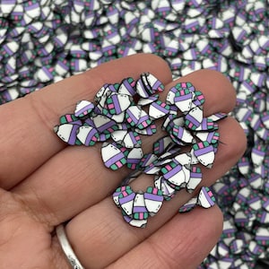 May include: A close-up of a hand holding a pile of small, white, purple, and green heart-shaped resin charms. The charms have a black outline and a geometric pattern.